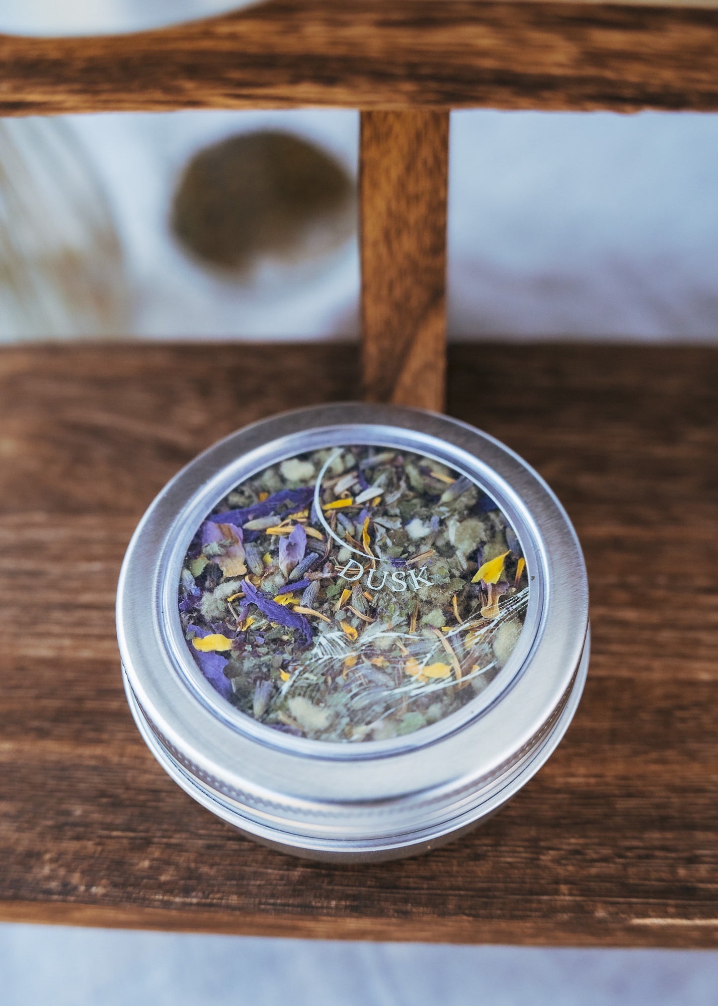 A product image of Ophelia’s DUSK Blend in a metal reusable tin on wooden surface. There is a clear window on the top of the tin showing blue lotus, mullein, tulsi, mugwort and lavender.